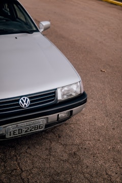A close-up view of the front portion of a silver Volkswagen car, parked on a paved surface. The vehicle's grille, headlight, and license plate are visible. The background consists of a textured asphalt road, with a small section of a yellow curb in the distance.