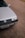 A close-up view of the front portion of a silver Volkswagen car, parked on a paved surface. The vehicle's grille, headlight, and license plate are visible. The background consists of a textured asphalt road, with a small section of a yellow curb in the distance.