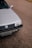 A close-up view of the front portion of a silver Volkswagen car, parked on a paved surface. The vehicle's grille, headlight, and license plate are visible. The background consists of a textured asphalt road, with a small section of a yellow curb in the distance.