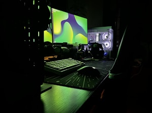 A dark, moody setup featuring a dual-monitor configuration on a desk. The monitors display abstract green shapes, casting an eerie glow. A mechanical keyboard and a gaming mouse sit on a large mouse pad. The translucent computer case reveals illuminated internal components, adding a futuristic touch.