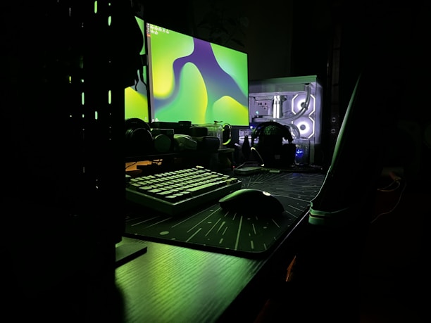 A dark, moody setup featuring a dual-monitor configuration on a desk. The monitors display abstract green shapes, casting an eerie glow. A mechanical keyboard and a gaming mouse sit on a large mouse pad. The translucent computer case reveals illuminated internal components, adding a futuristic touch.