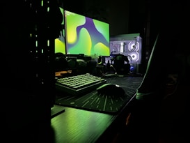 A dark, moody setup featuring a dual-monitor configuration on a desk. The monitors display abstract green shapes, casting an eerie glow. A mechanical keyboard and a gaming mouse sit on a large mouse pad. The translucent computer case reveals illuminated internal components, adding a futuristic touch.