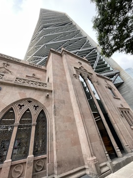 A juxtaposition of architectural styles is prominently displayed with a modern skyscraper featuring a unique geometric facade towering above a historic building with ornate stone carvings and arched windows. The contrast highlights the blend of contemporary and classic design elements.