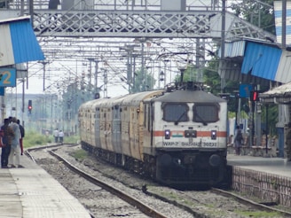 A train is arriving at a railway station with platform numbers 1 and 2 visible. There are people standing on the platform, a railway engine labeled 'WAP-7 GHAZIABAD 30232' is at the front of the train, and overhead wires and metal structures are present in the background.