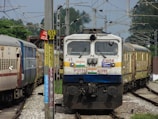 A train is approaching on a railway track with multiple coaches visible. Overhead electric lines and poles are present, and there are passengers looking out from the windows. The train is surrounded by signs and markers, and there is vegetation in the background.