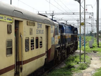 A train moves on railway tracks with a visible second class carriage. The yellow and cream-colored carriages have several windows, some with bars. A person stands at the door of one carriage, looking out. Overhead, electric wires and poles stretch across the sky. The tracks are surrounded by green grassy areas and some utility boxes on the side.
