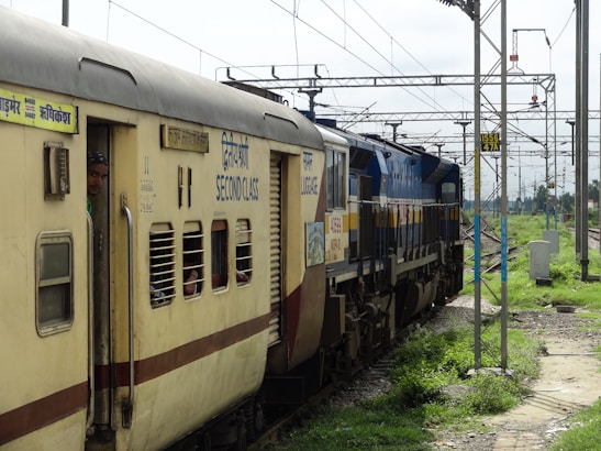 A train moves on railway tracks with a visible second class carriage. The yellow and cream-colored carriages have several windows, some with bars. A person stands at the door of one carriage, looking out. Overhead, electric wires and poles stretch across the sky. The tracks are surrounded by green grassy areas and some utility boxes on the side.