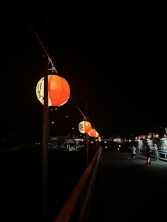 A series of illuminated red lanterns hang along a dark pathway at night. The lanterns create a warm, inviting glow against a shadowy background. A few people can be seen walking along the path, adding a touch of life to the otherwise tranquil scene.