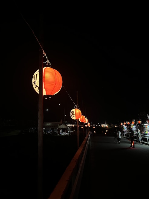 A series of illuminated red lanterns hang along a dark pathway at night. The lanterns create a warm, inviting glow against a shadowy background. A few people can be seen walking along the path, adding a touch of life to the otherwise tranquil scene.