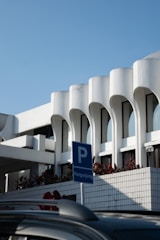 A modern architectural structure with white curvilinear features and large windows. The building is complemented by a clear blue sky and bordered with plants. In the foreground, there is a blue parking sign with the word 'Pengunjung', indicating visitor parking, and a portion of a dark-colored car roof.