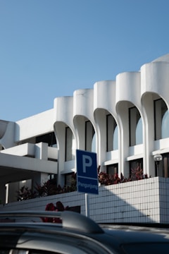 A modern architectural structure with white curvilinear features and large windows. The building is complemented by a clear blue sky and bordered with plants. In the foreground, there is a blue parking sign with the word 'Pengunjung', indicating visitor parking, and a portion of a dark-colored car roof.