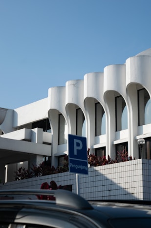 A modern architectural structure with white curvilinear features and large windows. The building is complemented by a clear blue sky and bordered with plants. In the foreground, there is a blue parking sign with the word 'Pengunjung', indicating visitor parking, and a portion of a dark-colored car roof.
