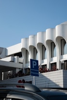 A modern architectural structure with white curvilinear features and large windows. The building is complemented by a clear blue sky and bordered with plants. In the foreground, there is a blue parking sign with the word 'Pengunjung', indicating visitor parking, and a portion of a dark-colored car roof.