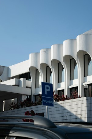 A modern architectural structure with white curvilinear features and large windows. The building is complemented by a clear blue sky and bordered with plants. In the foreground, there is a blue parking sign with the word 'Pengunjung', indicating visitor parking, and a portion of a dark-colored car roof.