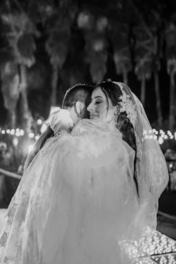 A couple is embracing in a romantic setting, likely during a wedding. The woman is wearing a bridal veil with floral decorations. The background is soft and features blurred palm trees and string lights, giving a dreamy atmosphere.