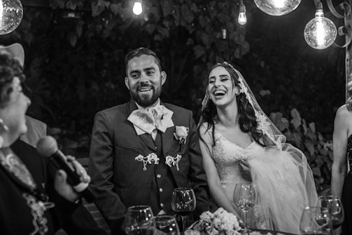 A joyful couple, consisting of a groom in a traditional suit and a bride in a wedding dress with a veil, is smiling and laughing. They are surrounded by friends, illuminated by hanging light bulbs, and have wine glasses in front of them. The background features dense foliage, contributing to a festive and intimate atmosphere.