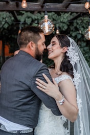 A joyous couple shares an intimate moment under hanging lights, with the woman wearing an elegant lace wedding dress and veil, and the man in a dark suit. They are both smiling warmly at each other.