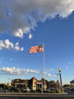 An American flag waves prominently against a blue sky with some clouds. Below, a McDonald's restaurant is visible along with several parked cars in the lot. The scene appears to be taken in daylight with bright conditions.