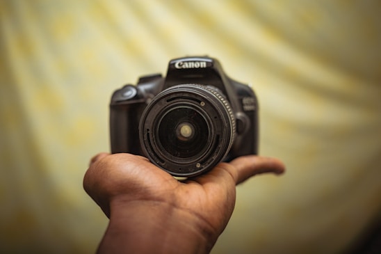 A person is holding a Canon camera, displayed prominently against a soft, blurred background.