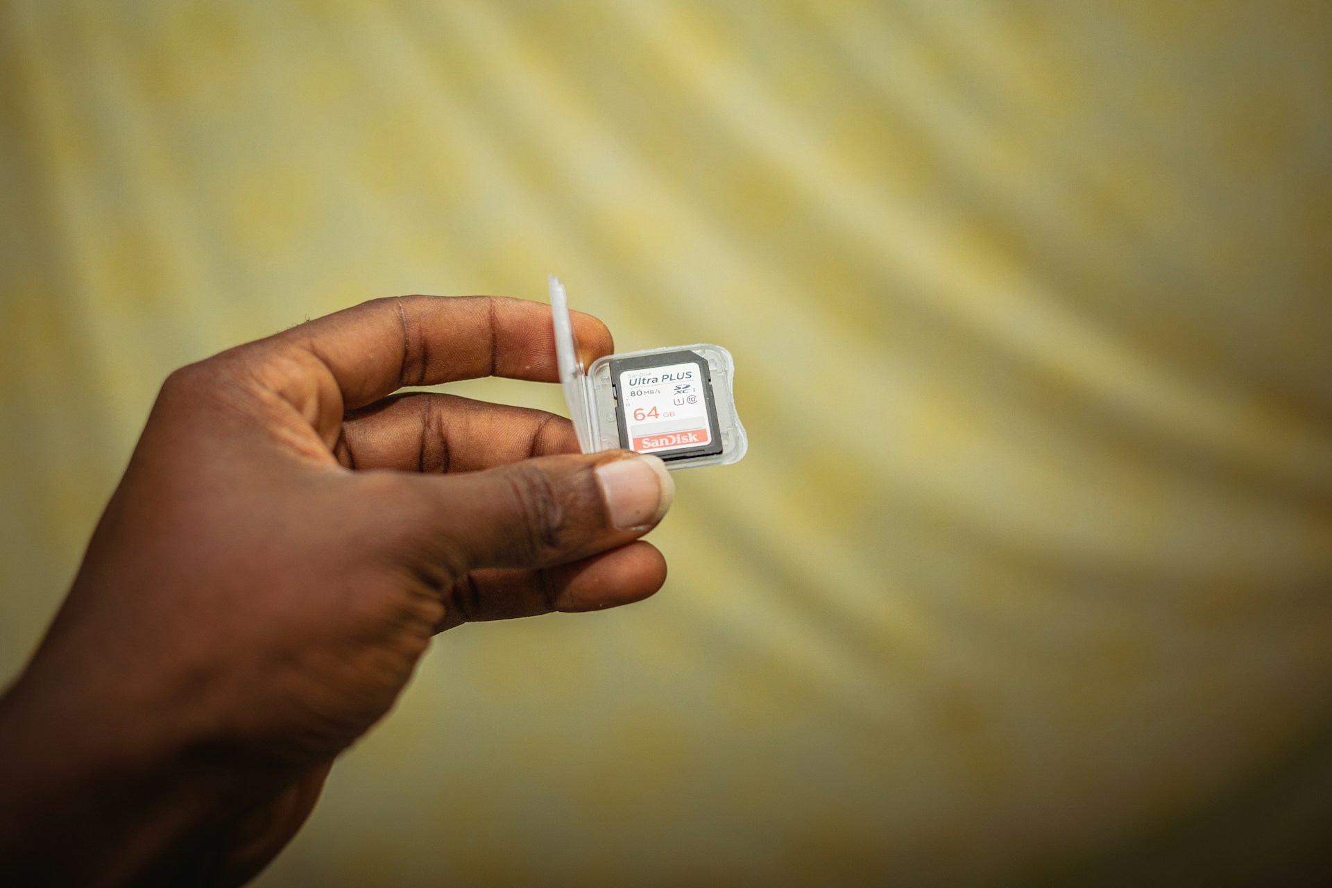 A hand is holding a SanDisk Ultra Plus 64GB memory card and its packaging against a softly blurred yellowish background. The fingers are delicately gripping the clear plastic case containing the card, which is labeled with red and white text.