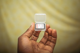 A hand holding a small clear plastic case containing a memory card. The card inside is labeled with the brand name and storage capacity details. The background is blurred and features a soft yellow tone.