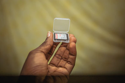 A hand holding a microSD card with a blurred laptop in the background.