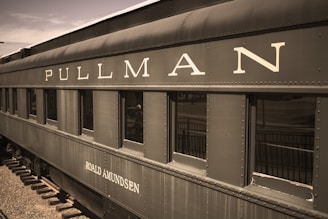 A vintage Pullman train car featuring riveted steel construction with large lettered signage reading 'PULLMAN' and 'ROALD AMUNDSEN'. The color is a faded, sepia tone, contributing to an antique appearance. The train car windows are dark, reflecting the surrounding environment, and the metal surface shows signs of wear and rust.