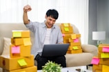 A person is sitting on a couch, animatedly raising a fist in the air with a smile, holding a laptop on their lap. Surrounding them are numerous cardboard boxes with colorful sticky notes attached, suggesting a home-based business setup.