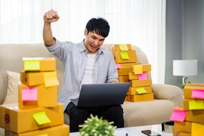 A person is sitting on a couch, animatedly raising a fist in the air with a smile, holding a laptop on their lap. Surrounding them are numerous cardboard boxes with colorful sticky notes attached, suggesting a home-based business setup.