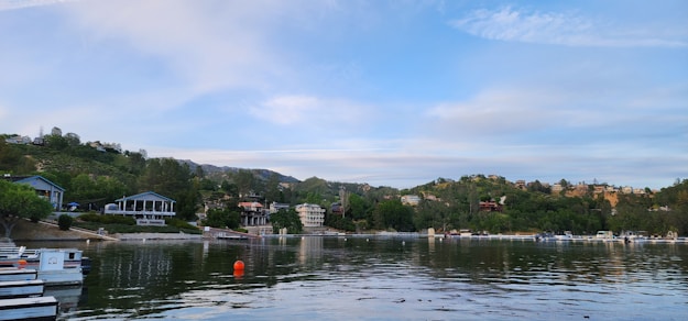 A calm lake is surrounded by a residential area with various houses and buildings. Some of the properties have docks extending into the water. The background features green, tree-covered hills. The sky is partly cloudy with a mix of blue and white clouds. There is a lone orange buoy floating in the lake.