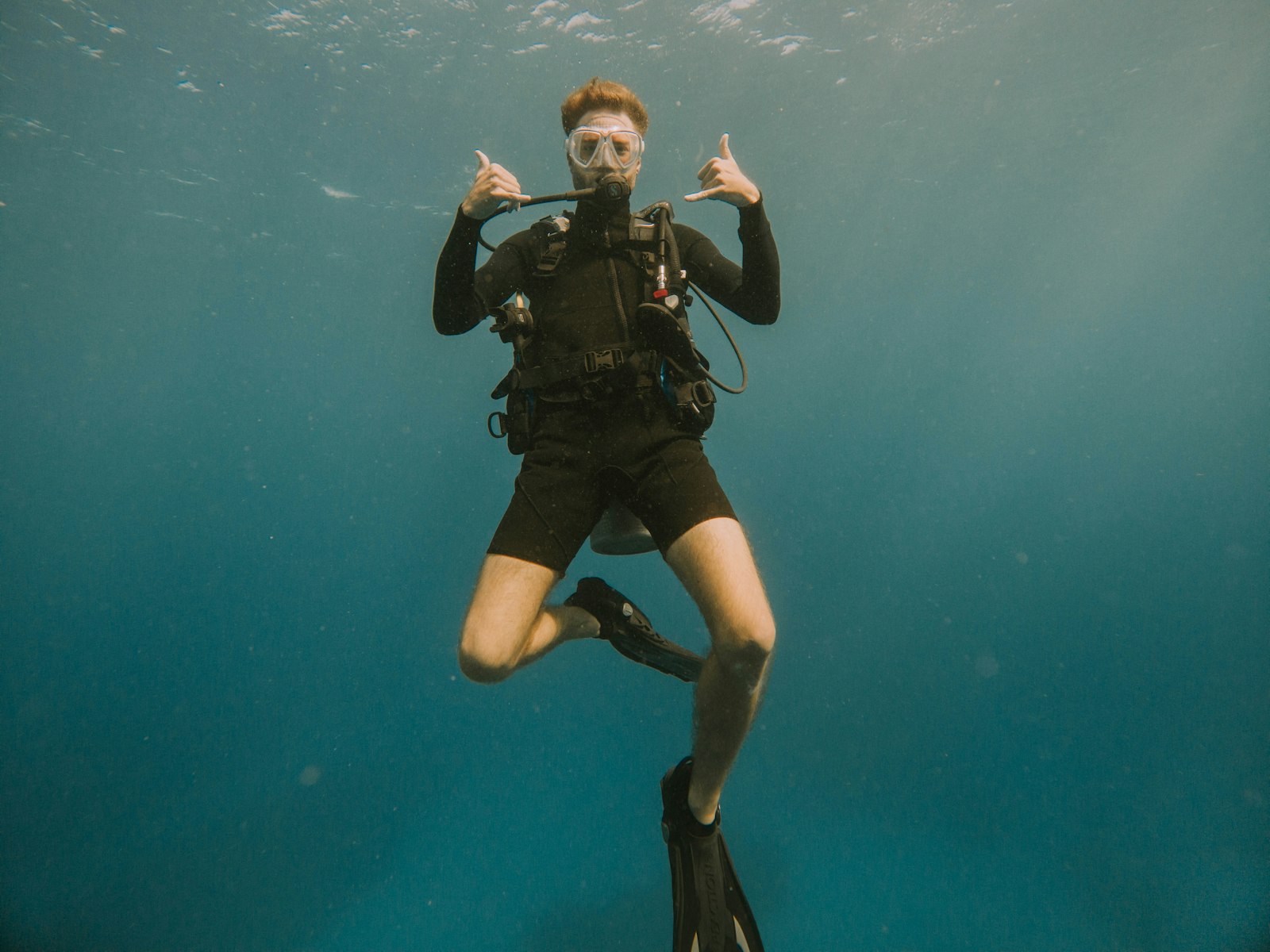 A scuba diver in black wetsuit and fins hovering in clear blue water, throwing a shaka sign toward the camera
