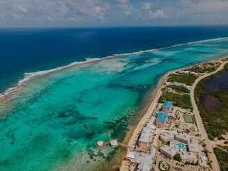 An aerial view of a coastal area with clear turquoise waters and coral reefs visible beneath the surface. A long strip of road runs parallel to the coast, with several buildings and resorts along the shoreline. The ocean stretches into the horizon.