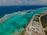 An aerial view of a coastal area with clear turquoise waters and coral reefs visible beneath the surface. A long strip of road runs parallel to the coast, with several buildings and resorts along the shoreline. The ocean stretches into the horizon.