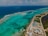 An aerial view of a coastal area with clear turquoise waters and coral reefs visible beneath the surface. A long strip of road runs parallel to the coast, with several buildings and resorts along the shoreline. The ocean stretches into the horizon.