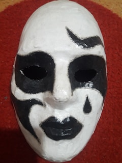 A white mask with black painted details, including stylized shapes around the eyes and a teardrop below one eye, is lying on a textured red surface. The mask has hollow eyes and black lips, creating a dramatic and enigmatic effect.