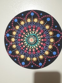 A circular, intricately patterned design resembling a mandala is composed of colorful dots arranged in symmetric patterns on a black background. The design features concentric circles with varying sizes and colors, including blues, reds, yellows, and greens. The symmetry and color arrangement create a visually pleasing and harmonious appearance.