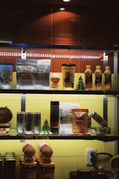 A shelf displays a variety of packaged snacks and drinks, including colorful cartoon-themed items and some beverages. The setting appears to be in a store or café, with bright lighting illuminating the products.