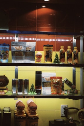 A shelf displays a variety of packaged snacks and drinks, including colorful cartoon-themed items and some beverages. The setting appears to be in a store or caf&eacute;, with bright lighting illuminating the products.