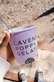 A hand holds a cup of lavender poppy gelato with visible purple gelato remnants inside. The container is predominantly lavender in color with bold black text. A spoon is inserted into the gelato. The background features a textured surface resembling bricks and stones, and a pair of sneakers can be seen below.