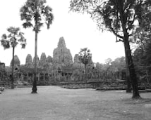 An ancient stone temple with intricate carvings is surrounded by slightly overgrown grass and tall palm trees. The temple architecture features multiple towers with detailed artistic elements, amidst a landscape that suggests a historical or archaeological site.