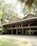 A traditional wooden building with a raised platform supported by wooden pillars stands amidst a lush green environment. The structure is adorned with multiple colorful flags and surrounded by tall trees, creating a serene and natural setting.