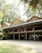 A traditional wooden building with a raised platform supported by wooden pillars stands amidst a lush green environment. The structure is adorned with multiple colorful flags and surrounded by tall trees, creating a serene and natural setting.