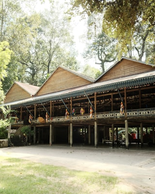 A traditional wooden building with a raised platform supported by wooden pillars stands amidst a lush green environment. The structure is adorned with multiple colorful flags and surrounded by tall trees, creating a serene and natural setting.