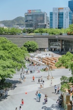 A lively urban plaza with multiple people walking and interacting in an open area. There are green trees surrounding the space, with a notable presence of buildings in the background, including a structure with colorful signage atop. The area seems to be hosting an outdoor event or market with booths and tents organized in the middle. Children can be seen playing, and adults are leisurely strolling around.