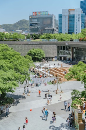 A lively urban plaza with multiple people walking and interacting in an open area. There are green trees surrounding the space, with a notable presence of buildings in the background, including a structure with colorful signage atop. The area seems to be hosting an outdoor event or market with booths and tents organized in the middle. Children can be seen playing, and adults are leisurely strolling around.
