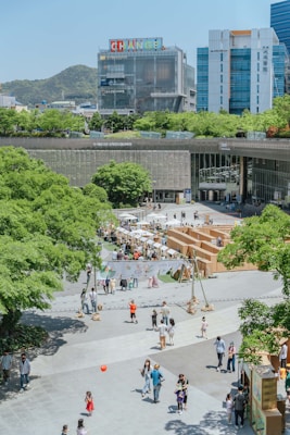 A lively urban plaza with multiple people walking and interacting in an open area. There are green trees surrounding the space, with a notable presence of buildings in the background, including a structure with colorful signage atop. The area seems to be hosting an outdoor event or market with booths and tents organized in the middle. Children can be seen playing, and adults are leisurely strolling around.
