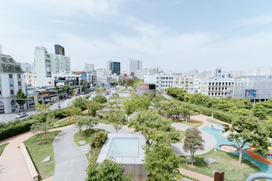 An urban park with lush green trees and pathways is surrounded by a cityscape of modern buildings and high-rises. The sky is clear, and the park features a playground area with colorful ground designs. Pedestrians and a few vehicles are visible on the adjacent streets.