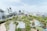 An urban park with lush green trees and pathways is surrounded by a cityscape of modern buildings and high-rises. The sky is clear, and the park features a playground area with colorful ground designs. Pedestrians and a few vehicles are visible on the adjacent streets.