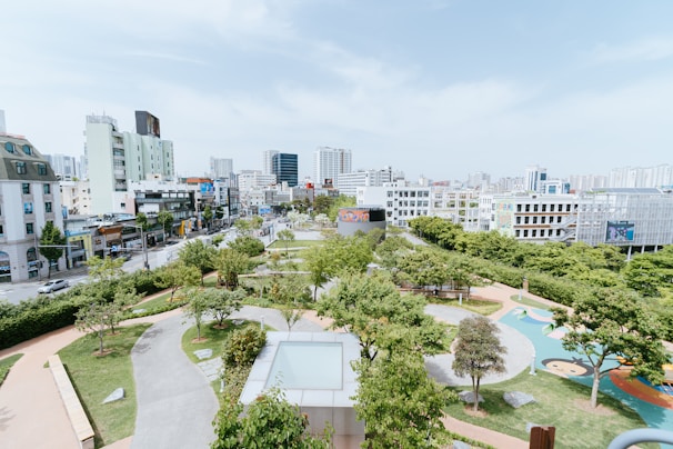 An urban park with lush green trees and pathways is surrounded by a cityscape of modern buildings and high-rises. The sky is clear, and the park features a playground area with colorful ground designs. Pedestrians and a few vehicles are visible on the adjacent streets.