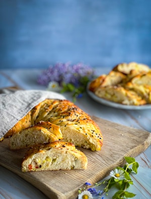 A braided loaf of bread with a golden-brown crust is partially sliced on a wooden cutting board. The bread is sprinkled with black sesame seeds and surrounded by fresh flowers and herbs. A plate with more bread slices is in the background against a soft blue backdrop.